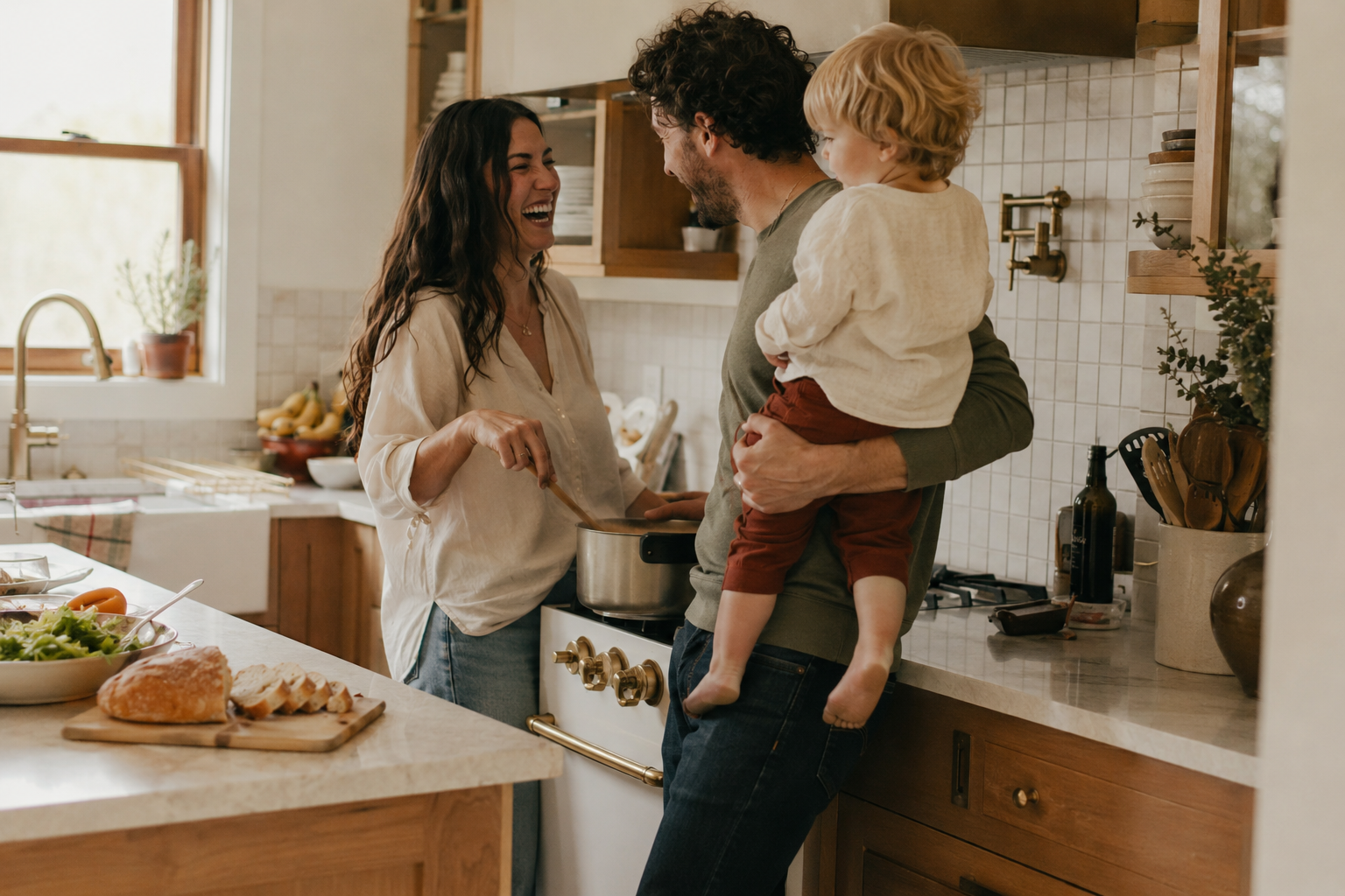A couple laughing together in a warm kitchen, one holding a toddler on their hip